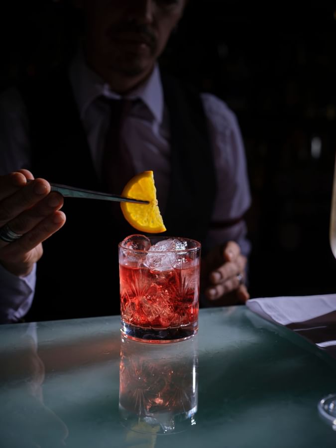 Red cocktail with ice on a table by a bartender holding an orange garnish in The Capital Bar, Tom Brown at The Capital