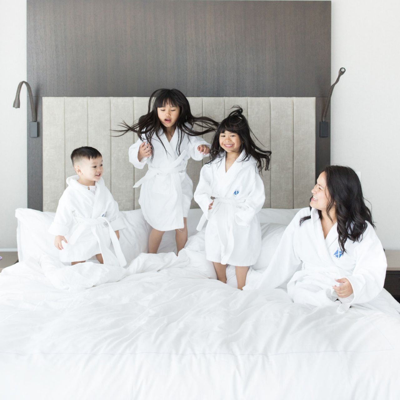 Mother and three children in white robes enjoying playful family time together on a large white bed at Hotel X Toronto