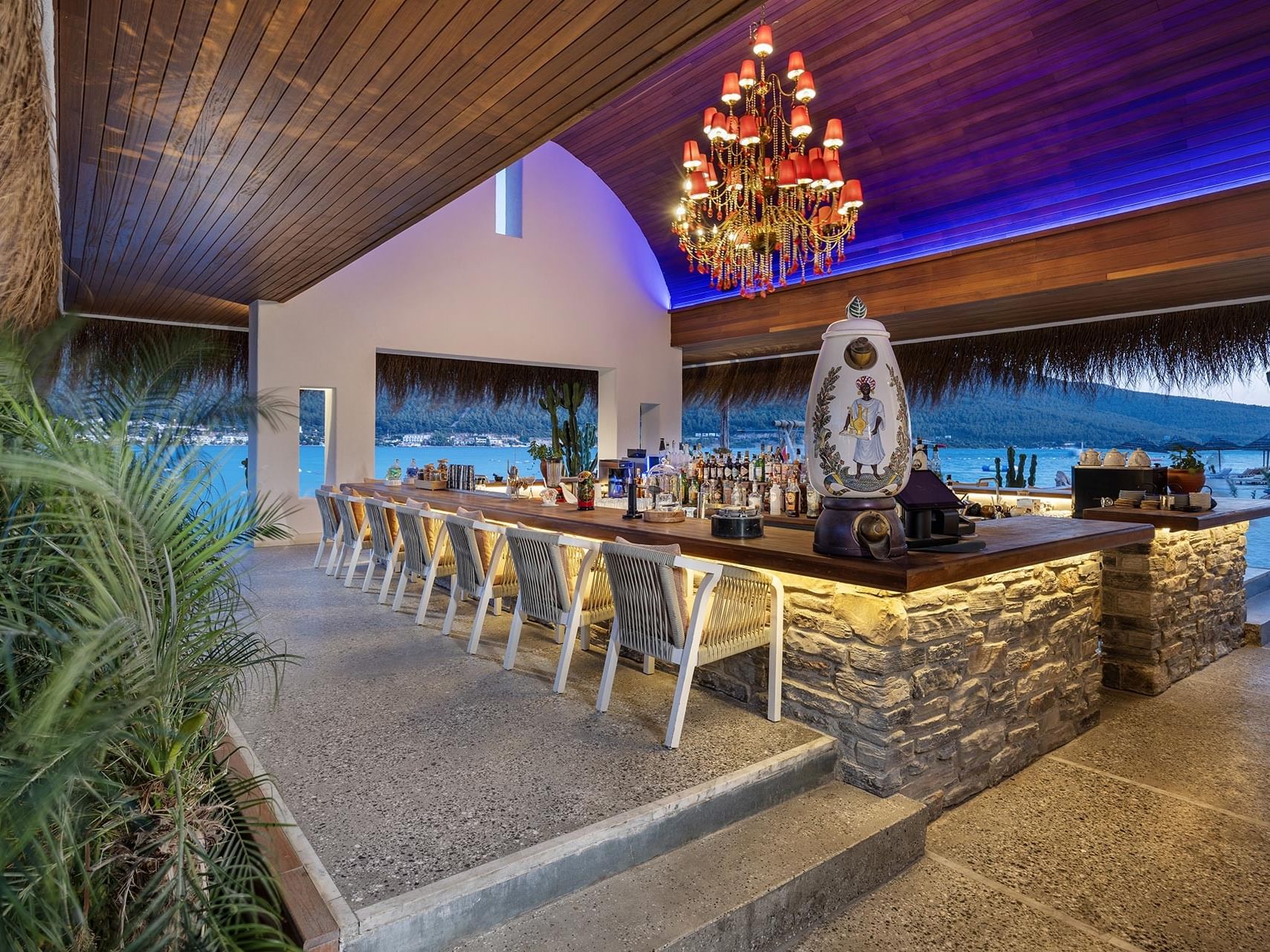 Coastal bar counter with chandeliers, a lit ceiling, and seating at Titanic Luxury Collection Bodrum.