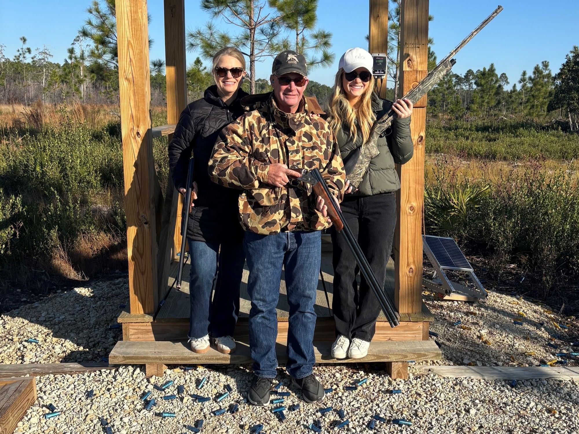Three people standing on a platform, holding shotguns, smiling for a photo in a natural setting.