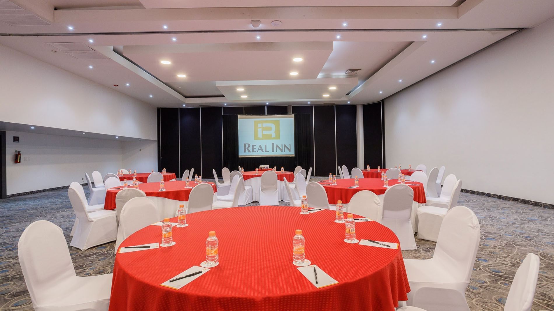 Banquets with red tablecloths and white chairs, featuring a projector screen in Florencia Hall at Real Inn Tijuana