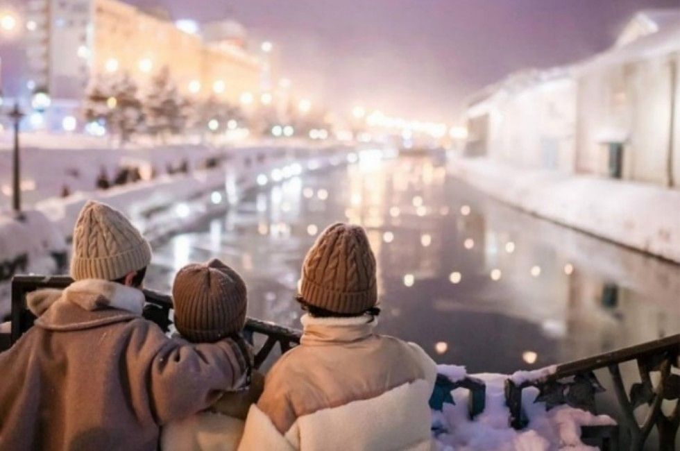 Three people in winter coats standing on bridge overlooking illuminated Otaru Canal near Grand Park Otaru