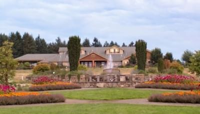 A garden with colorful flowers, a central fountain, and oregon garden resort in the background under a cloudy sky.