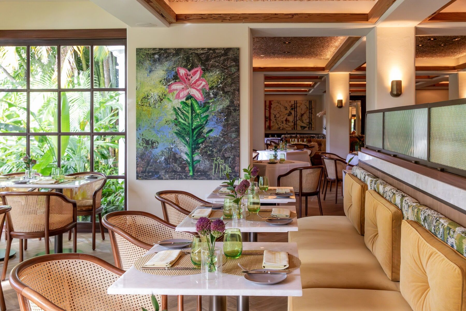 An elegant restaurant interior with set tables, cushioned seating, and a large floral painting on the wall.