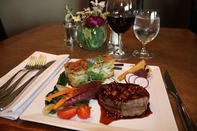 Steak dinner served on a plate with wine glasses set behind a flower vase at Hilltop Inn Salmon Arm