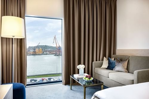 Hotel room with couch, coffee table, floor lamp, and large window overlooking a body of water and cityscape.