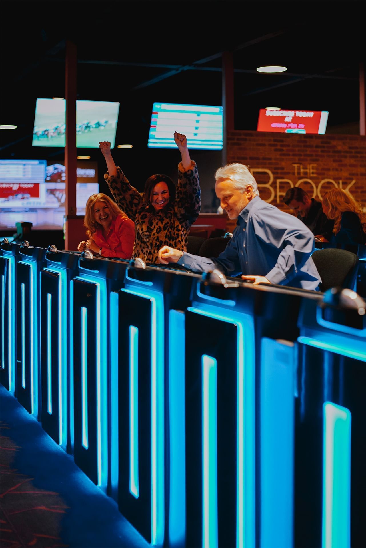 Guests celebrating behind blue-lit gaming terminals under digital betting screens in the casino area at The Brook