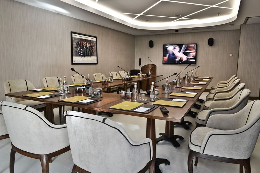 Grey chairs by a wooden table surrounding a large TV screen under bright lights in Kugu Hall at Warwick Ankara