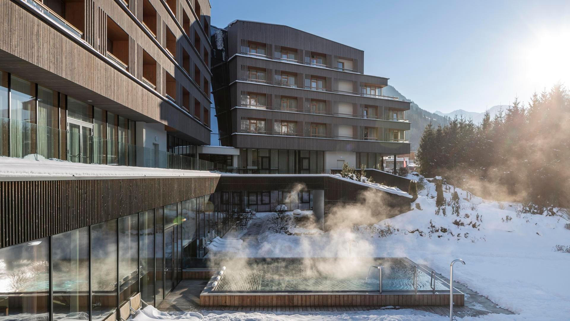 Modern hotel with steam-emitting hot tub in snowy outdoor area, surrounded by trees and mountains.