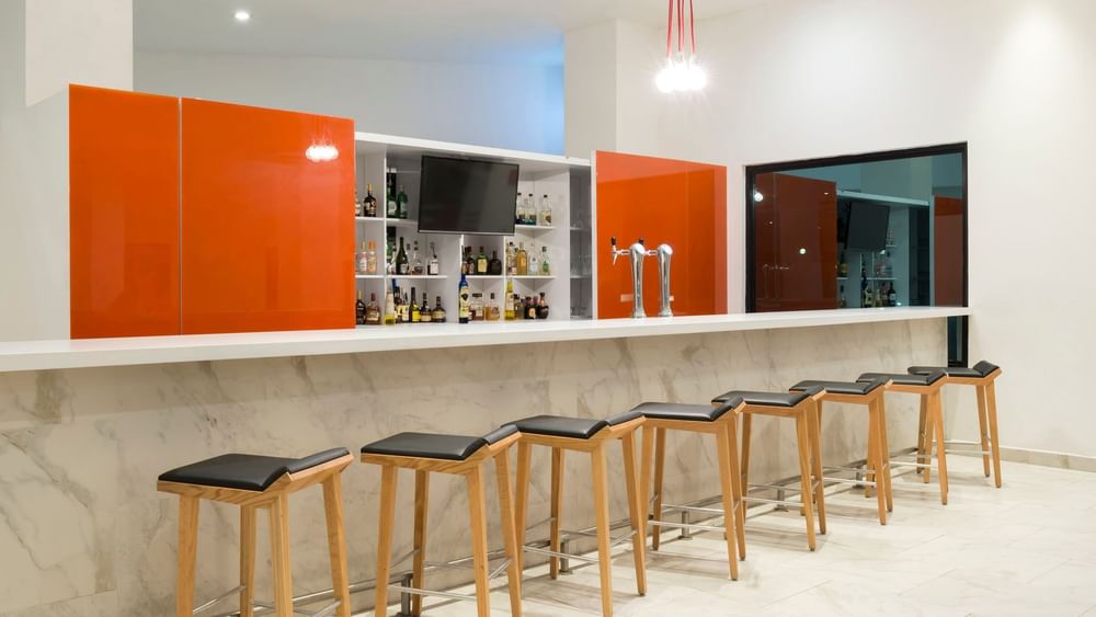 Black bar stools by a marble counter under bright lights in front of orange panels at the lobby bar in Real Inn Mexicali