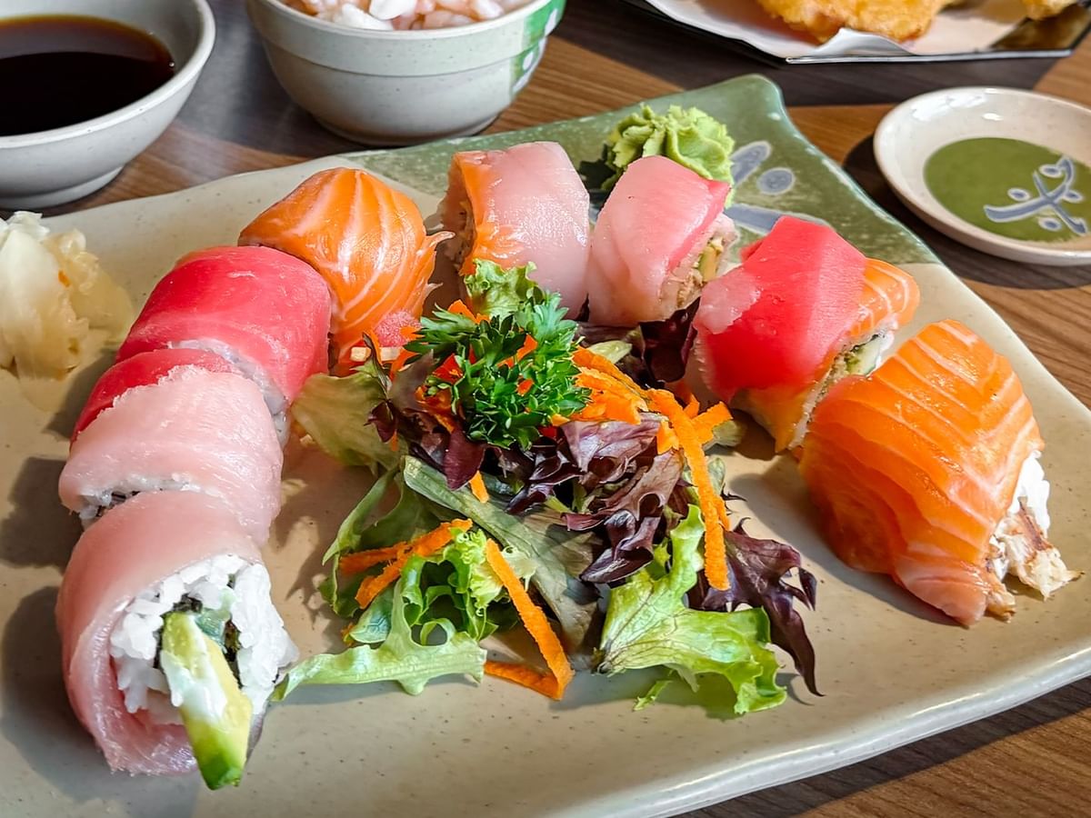 Plate of sushi rolls, sashimi, and salad with side dishes and dipping sauce on a wooden table.