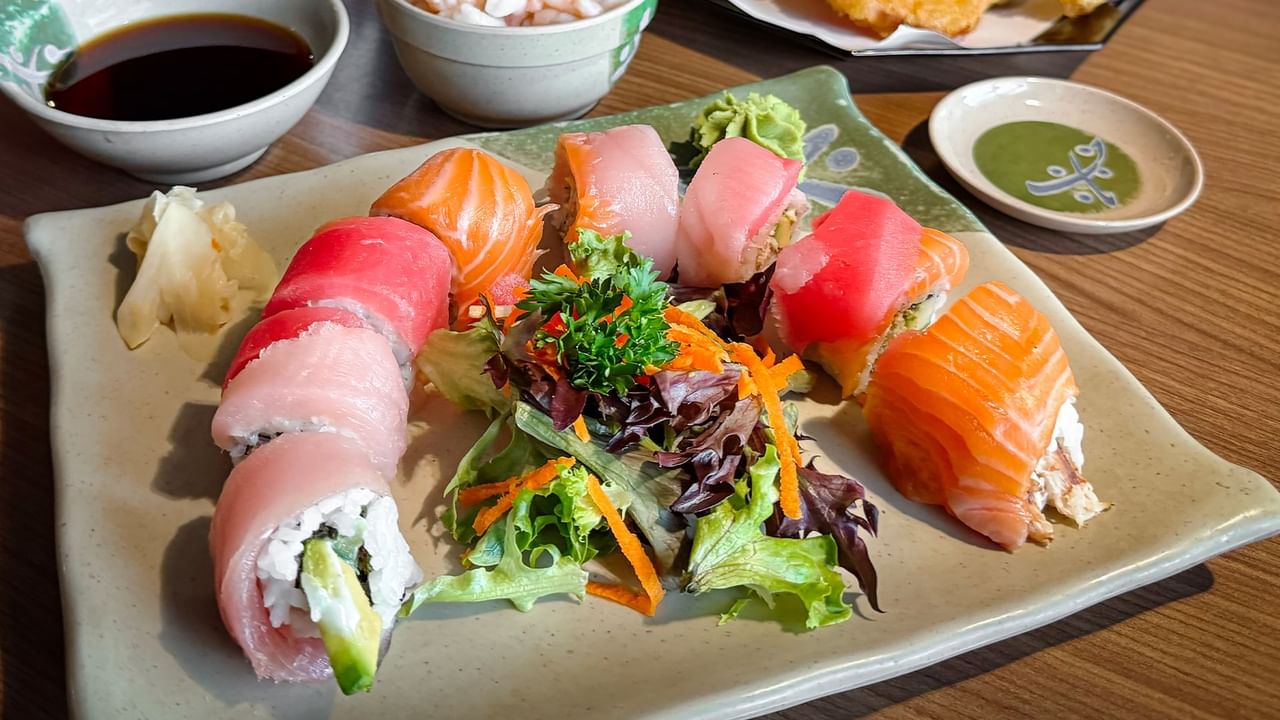 Plate of sushi rolls, sashimi, and salad with side dishes and dipping sauce on a wooden table.