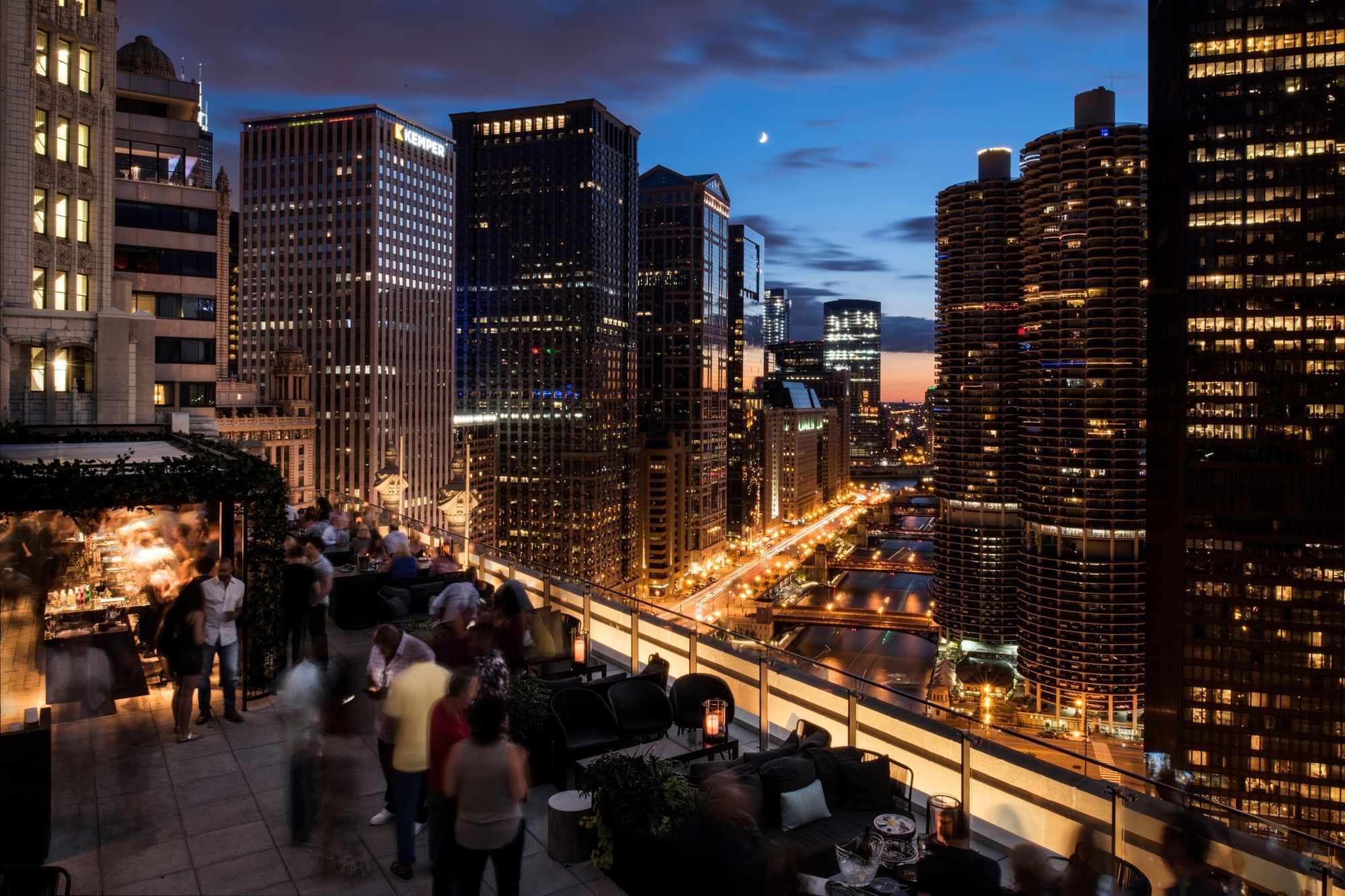A rooftop bar with people overlooking a city skyline at dusk.