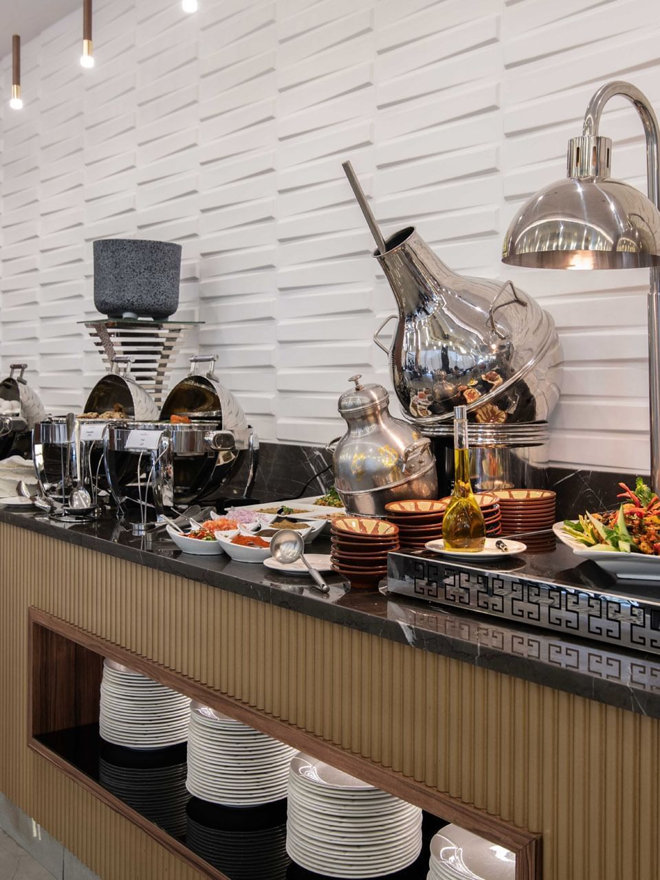 Buffet line with silver chafing dishes by a white textured wall in La Maison at Warwick Al Jubail