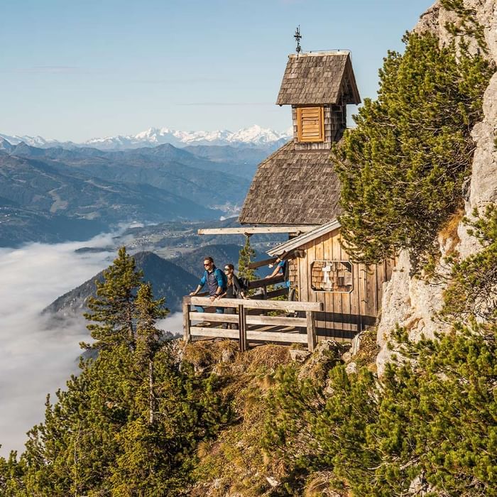 Coppia di escursionisti presso una capanna in legno su una scogliera con vista sulle montagne.