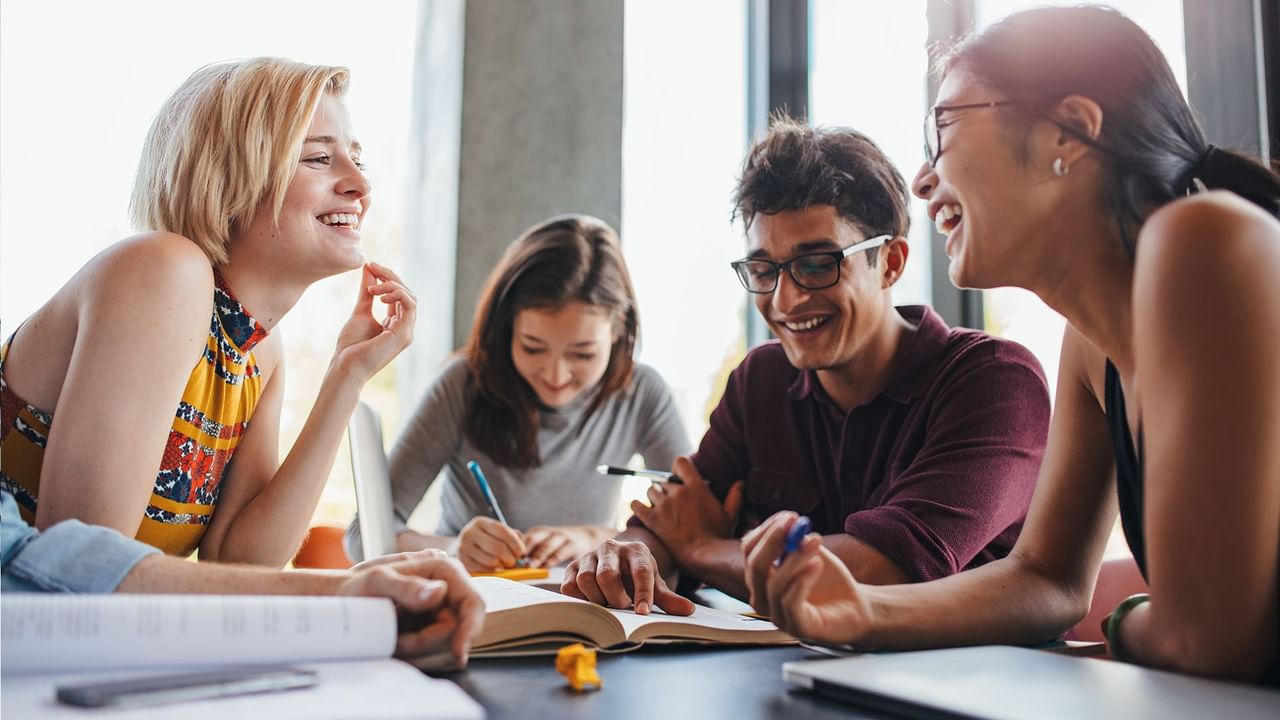 A group of students are sitting around a table, laughing and writing in notebooks.