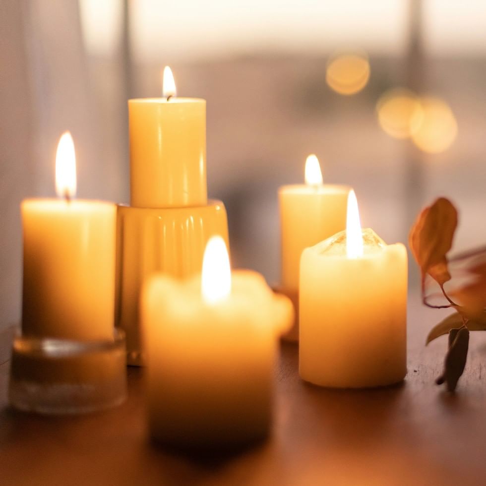 Five lit candles on a table with blurred window and lights in the background.