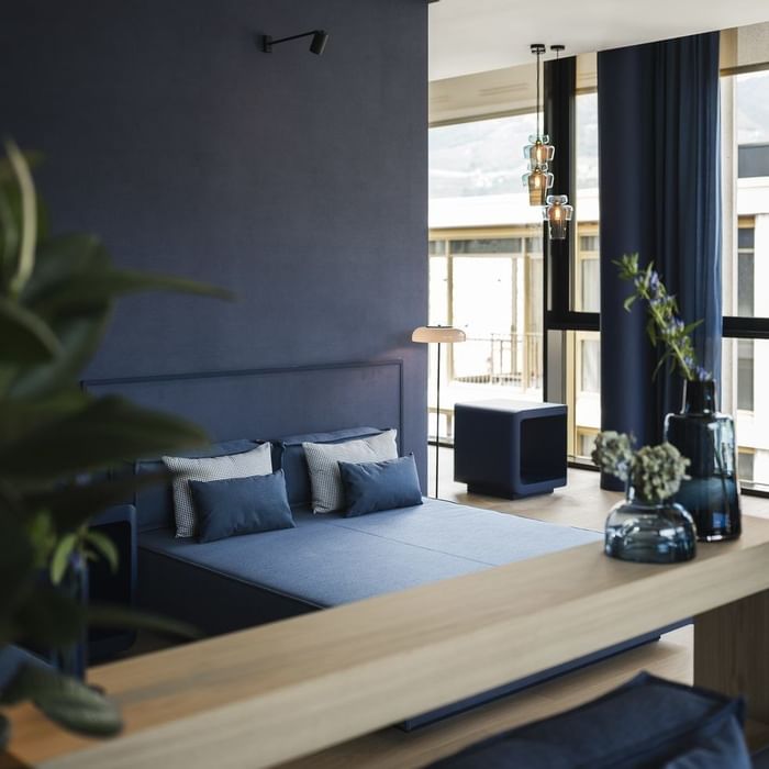 Blauer Bettbereich mit blauen Vorhängen, Pflanzen, Tisch und Fenstern im Hintergrund.
