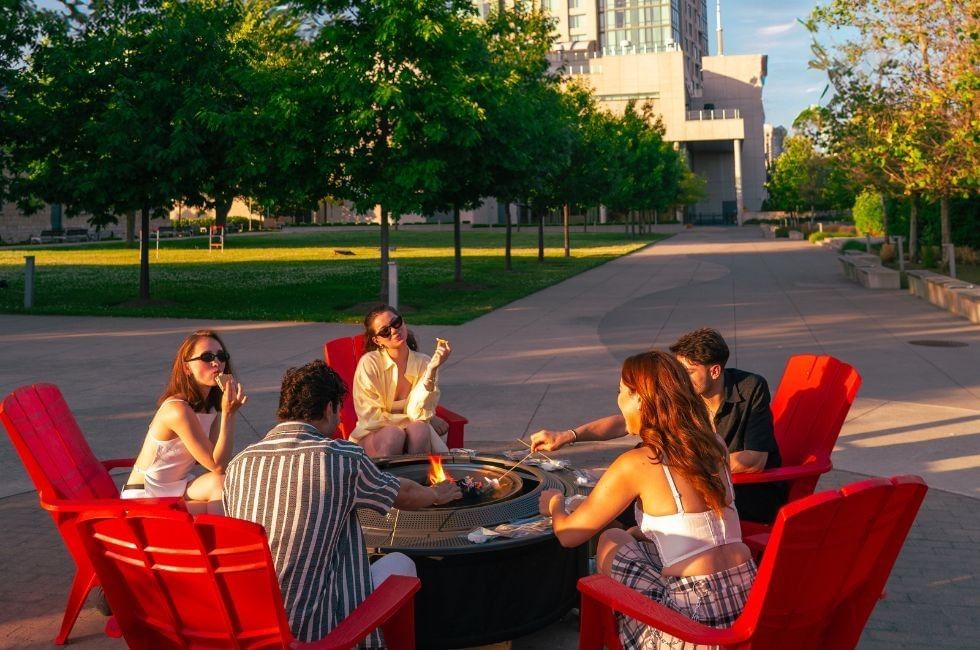 Friends seated on red chairs surrounding a fire pit by green trees under warm evening sunlight at Hotel X Toronto