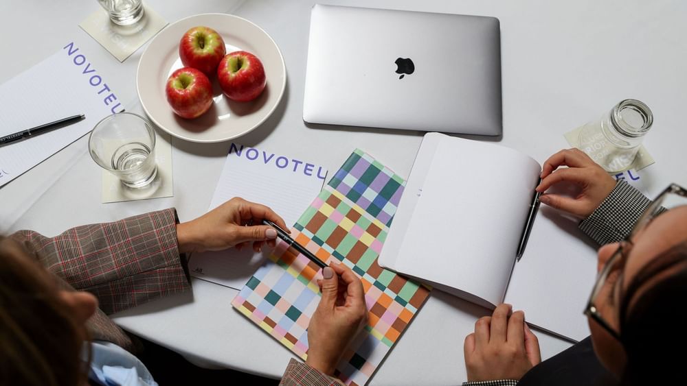 Aerial view of two ladies at a Freshwater Ballroom table with a laptop, notebooks, pens & apples at Novotel Olympic Park