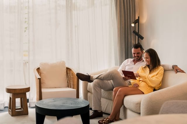 Happy couple on a cozy cream sofa by a wicker armchair under soft light in a luxury suite at Elysium Manly hotel