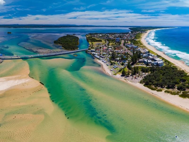 Aerial view of a coastal town with a bridge over a turquoise inlet, adjacent to a sandy beach near Mercure Kooindah Waters