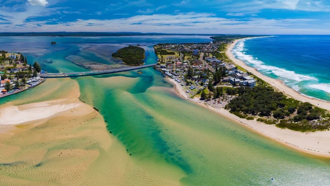 Aerial view of a coastal town with a bridge over a turquoise inlet, adjacent to a sandy beach near Mercure Kooindah Waters