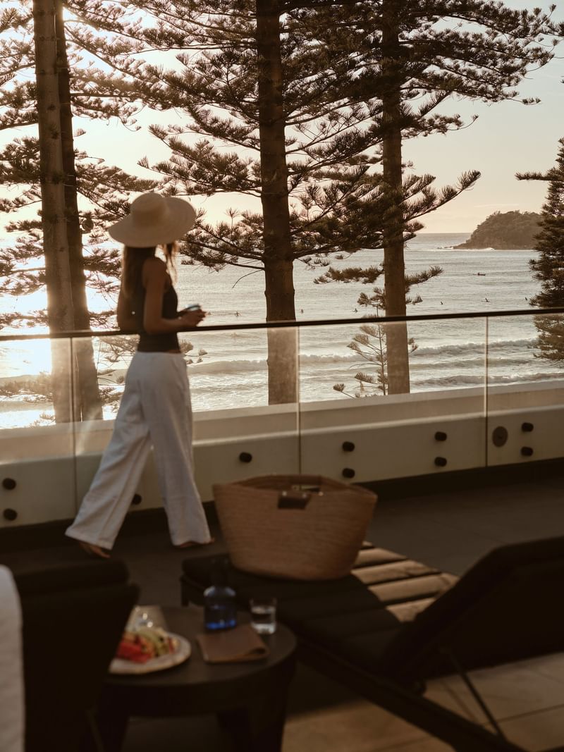 Woman at Elysium Manly walks by a glass balcony railing to look at the ocean waves, under a warm sunset, best hotels in Manly