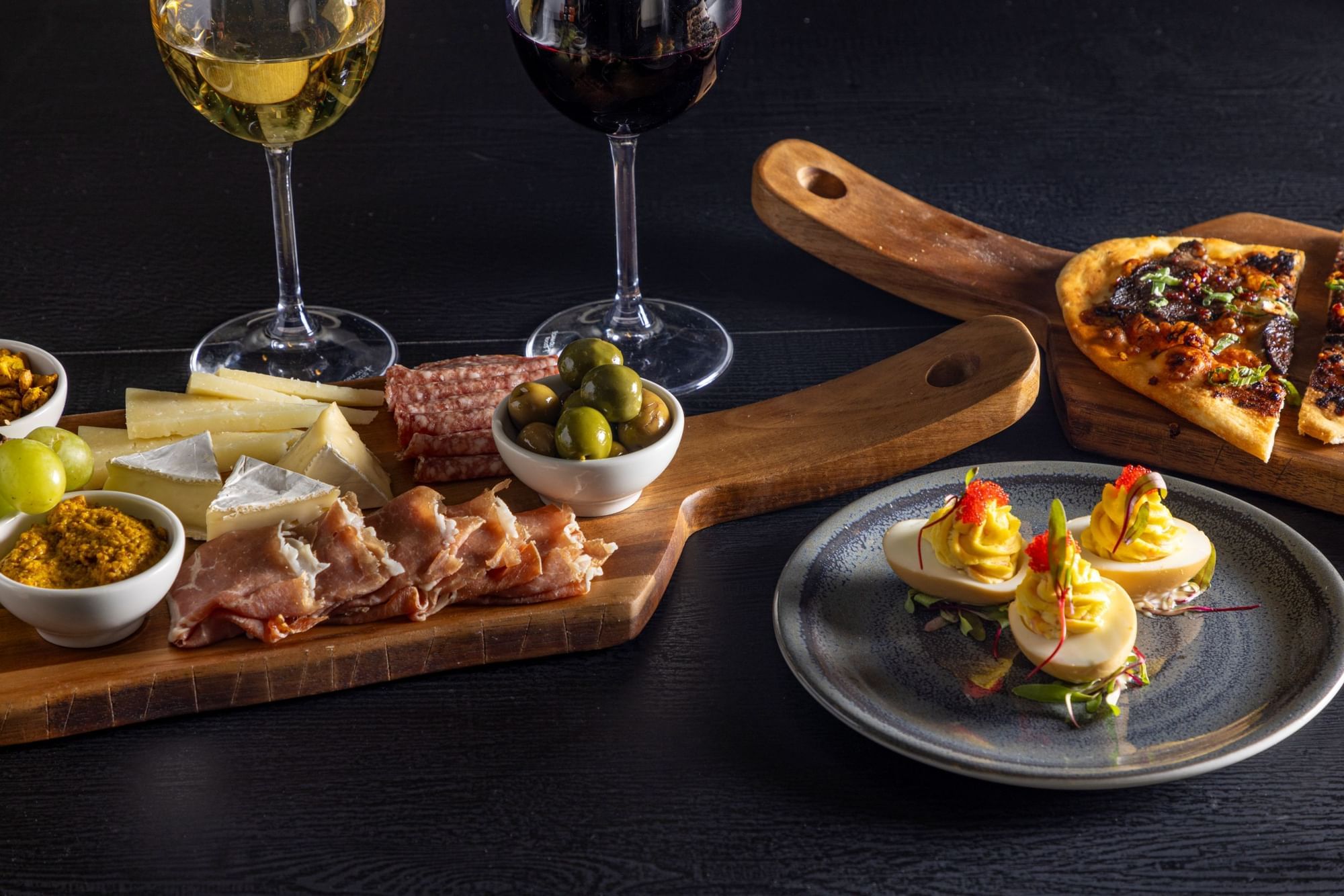 A selection of food on a table including a charcuterie board with cheese, meat, and olives, a plate of deviled eggs, and a pizza.