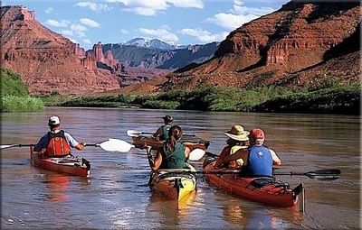 A group of kayakers paddle on the Colorado River, surrounded by rock cliffs under a cloudy sky near Moab Valley Inn