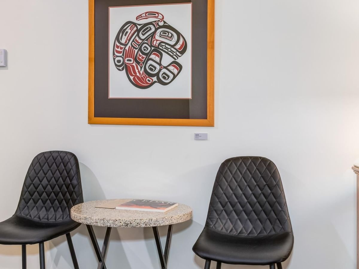 Two chairs and a small table with a book, near a framed Indigenous art.