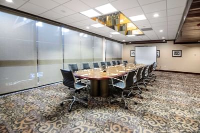 Long conference tables with black swivel chairs facing a large projector screen in the Meeting Room at Miami Lakes Hotel