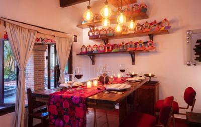 A wooden dining table set with candles, glasses, and red chairs in a room with decorative shelves and curtains.