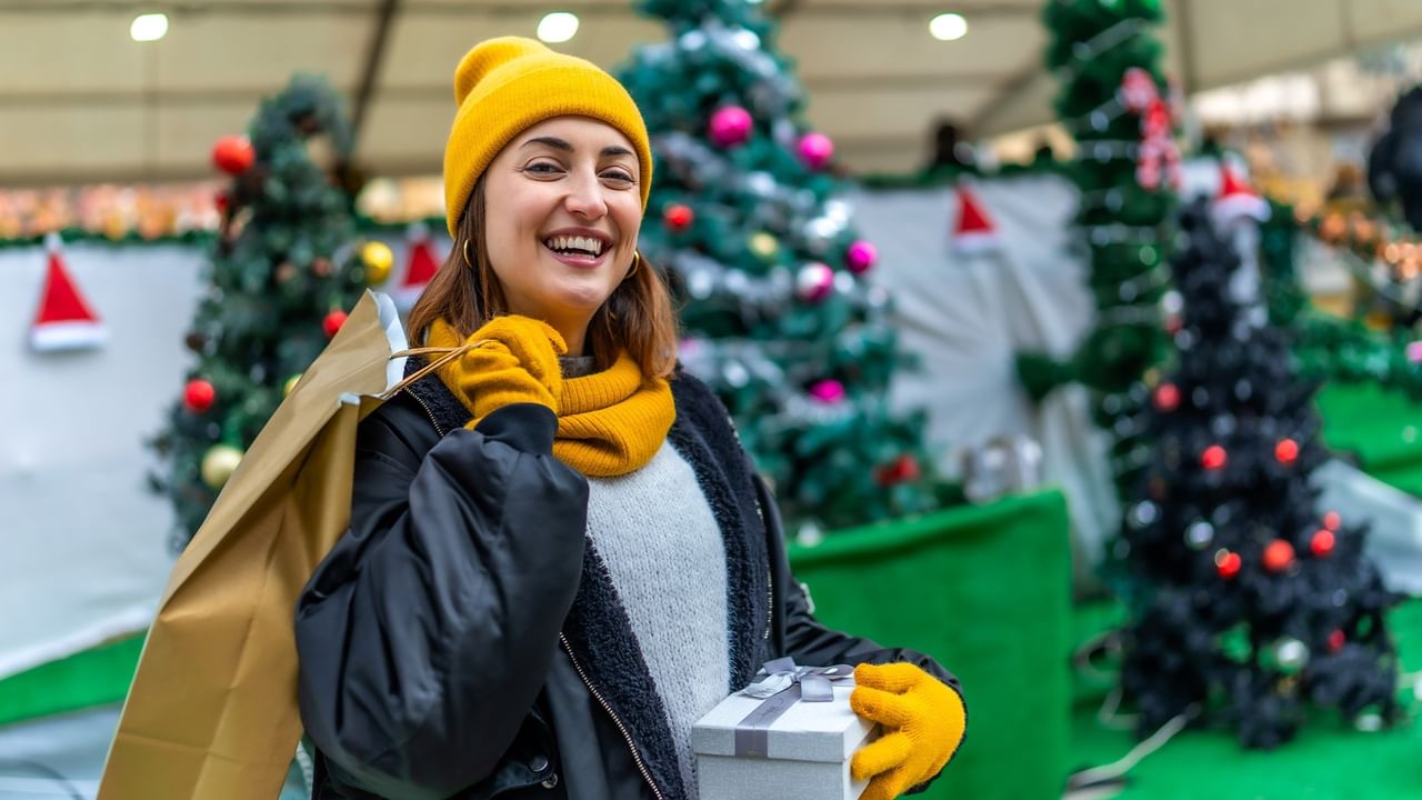 Smiling woman in yellow hat and scarf holding shopping bags and gift box at Christmas Gifts Expo with decorated trees.
