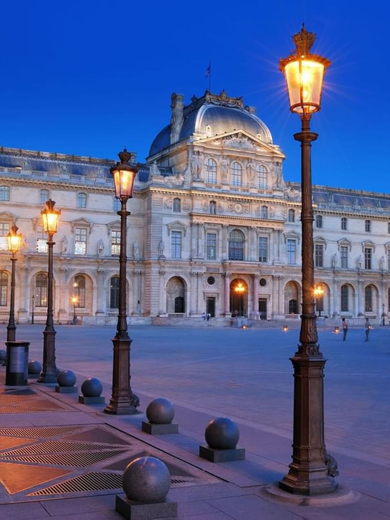 The Louvre Palace by glowing street lamps under a dark blue evening near Warwick Paris Champs Elysées