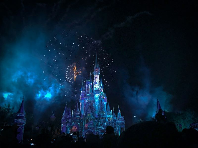 The image shows Cinderella Castle at night, illuminated with blue and purple lights. Fireworks are exploding in the sky above the castle. People appear to be watching in the foreground.