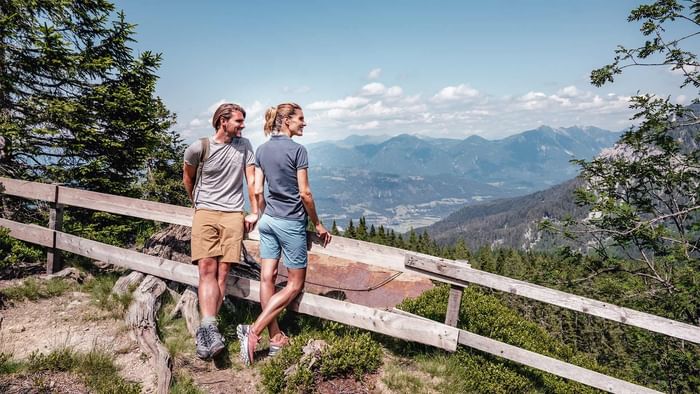 Couple enjoying a mountain view from a wooden fence near Falkensteiner Hotel Sonnenalpe