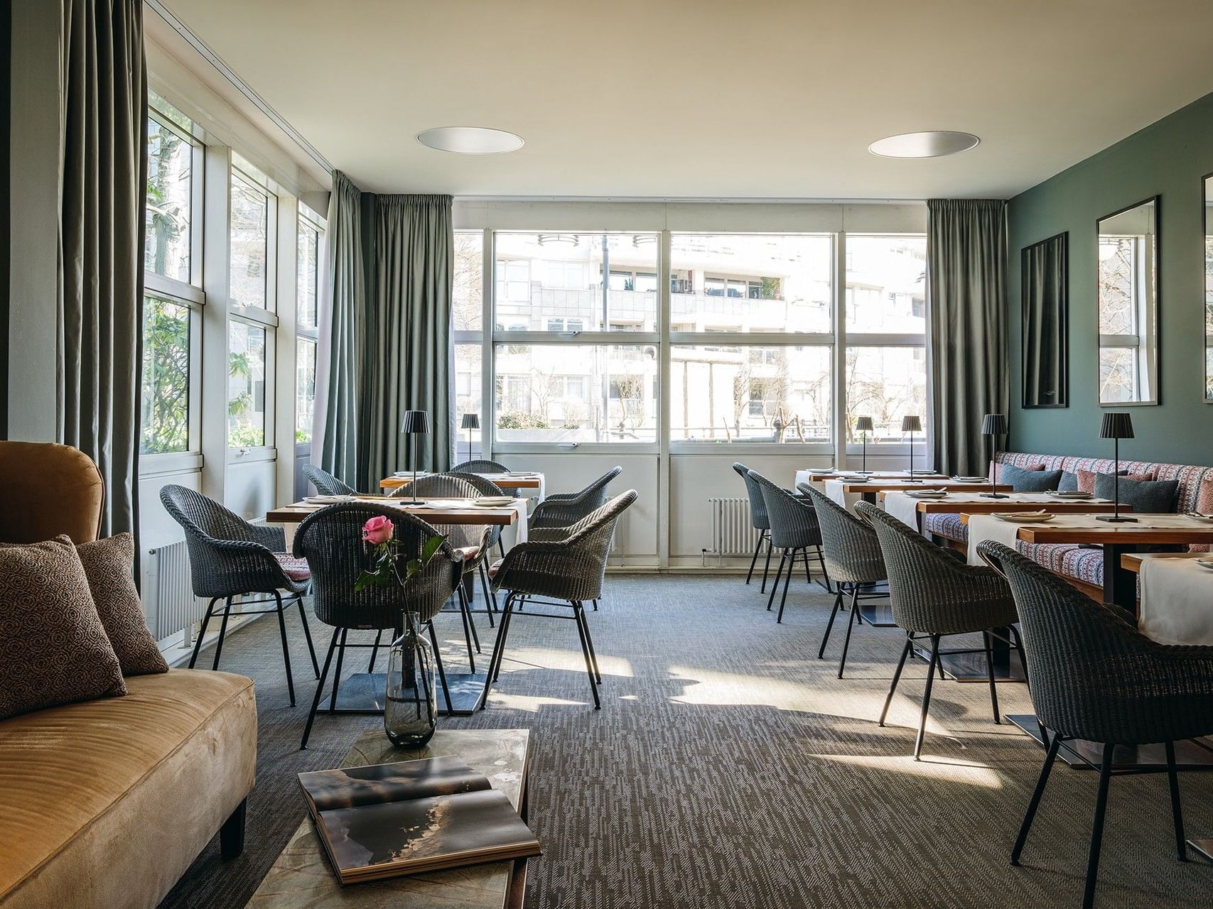 Ein Essbereich mit Tischen, Stühlen, einer Couch und großen Fenstern, die natürliches Licht hereinlassen.