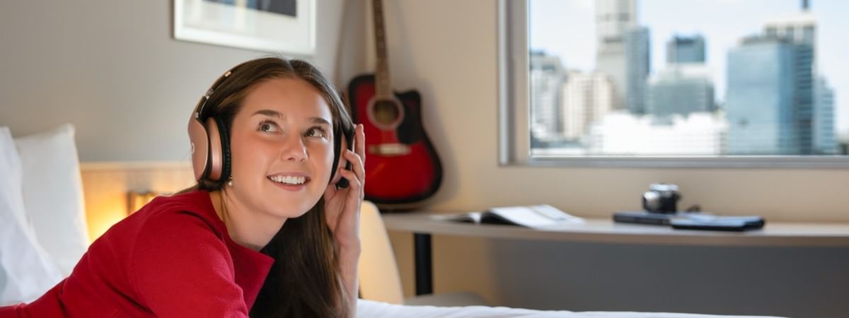 Guest relaxing in headphones on a bed at ibis Sydney Darling Harbour, with a Sydney skyline view.
