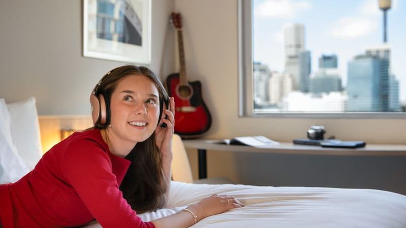 Guest relaxing in headphones on a bed at ibis Sydney Darling Harbour, with a Sydney skyline view.