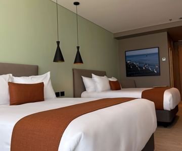 Two beds with orange accent throws under pendant lights and seabird art in a spacious guest room at Hotel B3 Sibelius