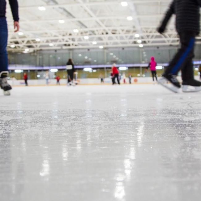 Skaters glide across a busy indoor ice rink with bright overhead lighting and colourful visitors in the background