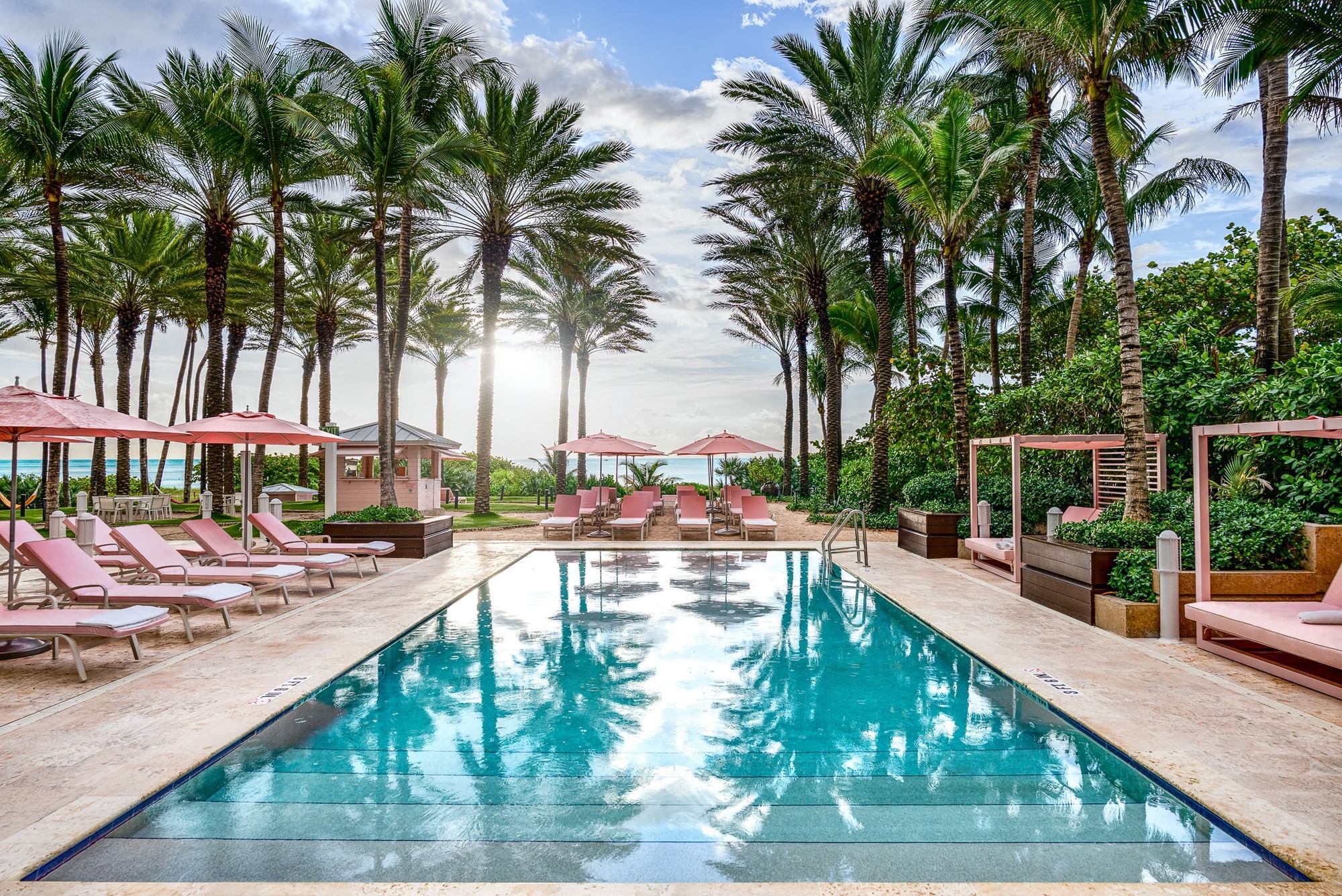 A serene poolside scene with a rectangular pool, surrounded by pink lounge chairs and umbrellas, palm trees, and a view of the ocean in the background.