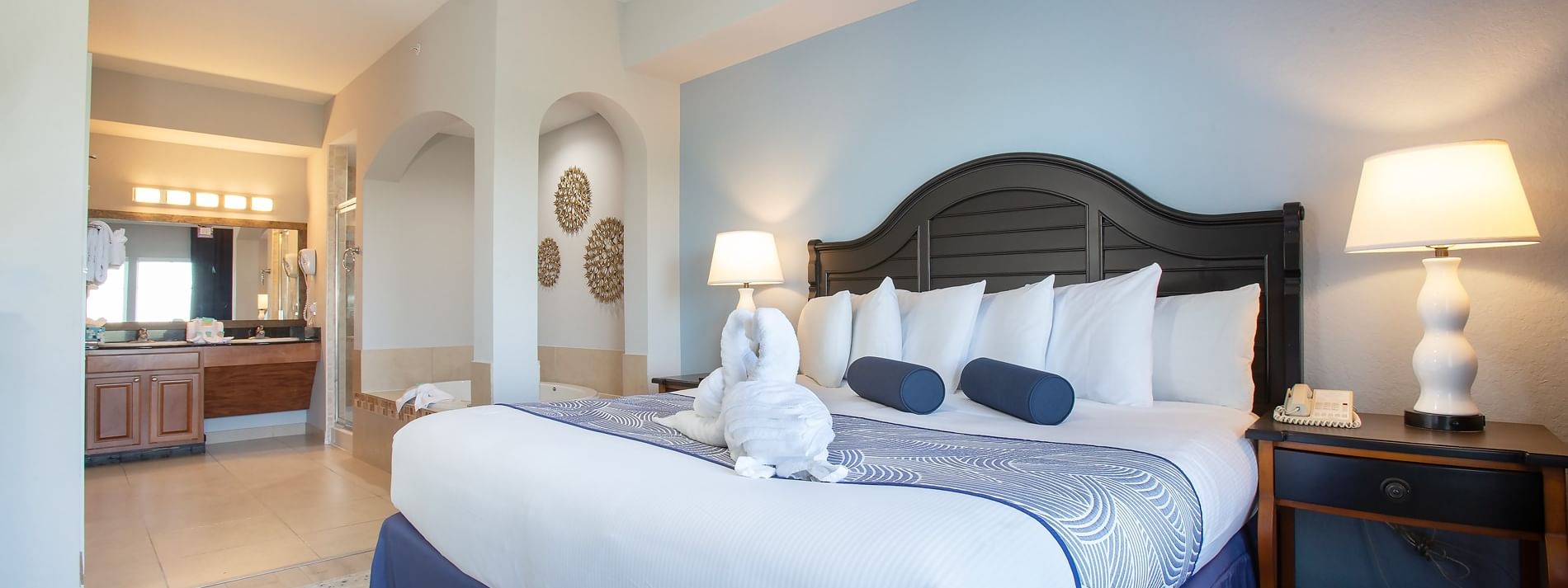 A wide-angle view of a hotel room with a king-sized bed, nightstand, and lamp, with a bathroom visible in the background.