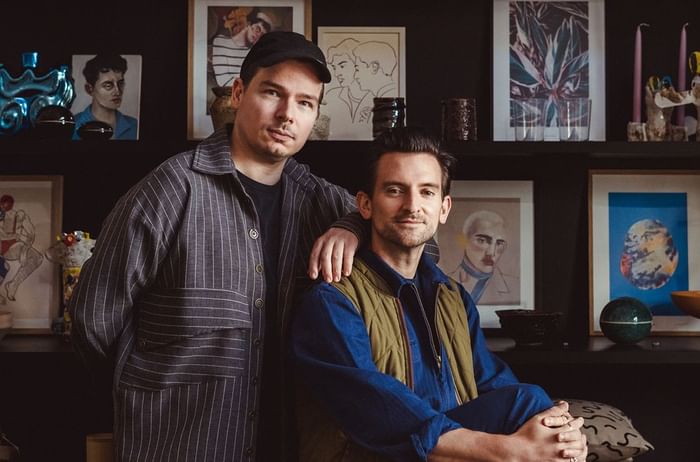 Two men pose together against a dark wall adorned with framed artworks at Hotel Motto Vienna