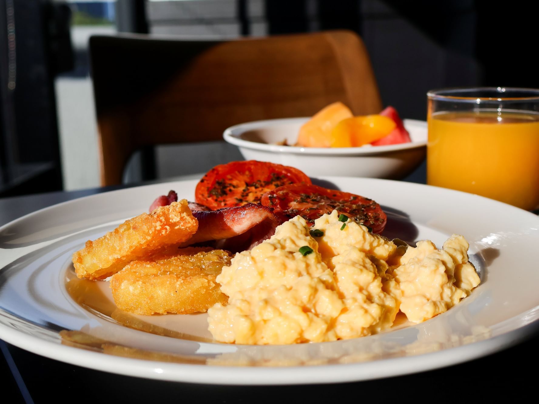 A hot breakfast plate featuring scrambled eggs, crispy hash browns, bacon, and grilled tomatoes, served with fresh orange juice and fruit at Frescos Restaurant.