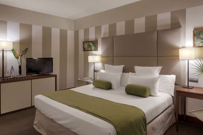 Bedroom with white bed, green blanket, white pillows, nightstands, TV, lamps, and striped walls.