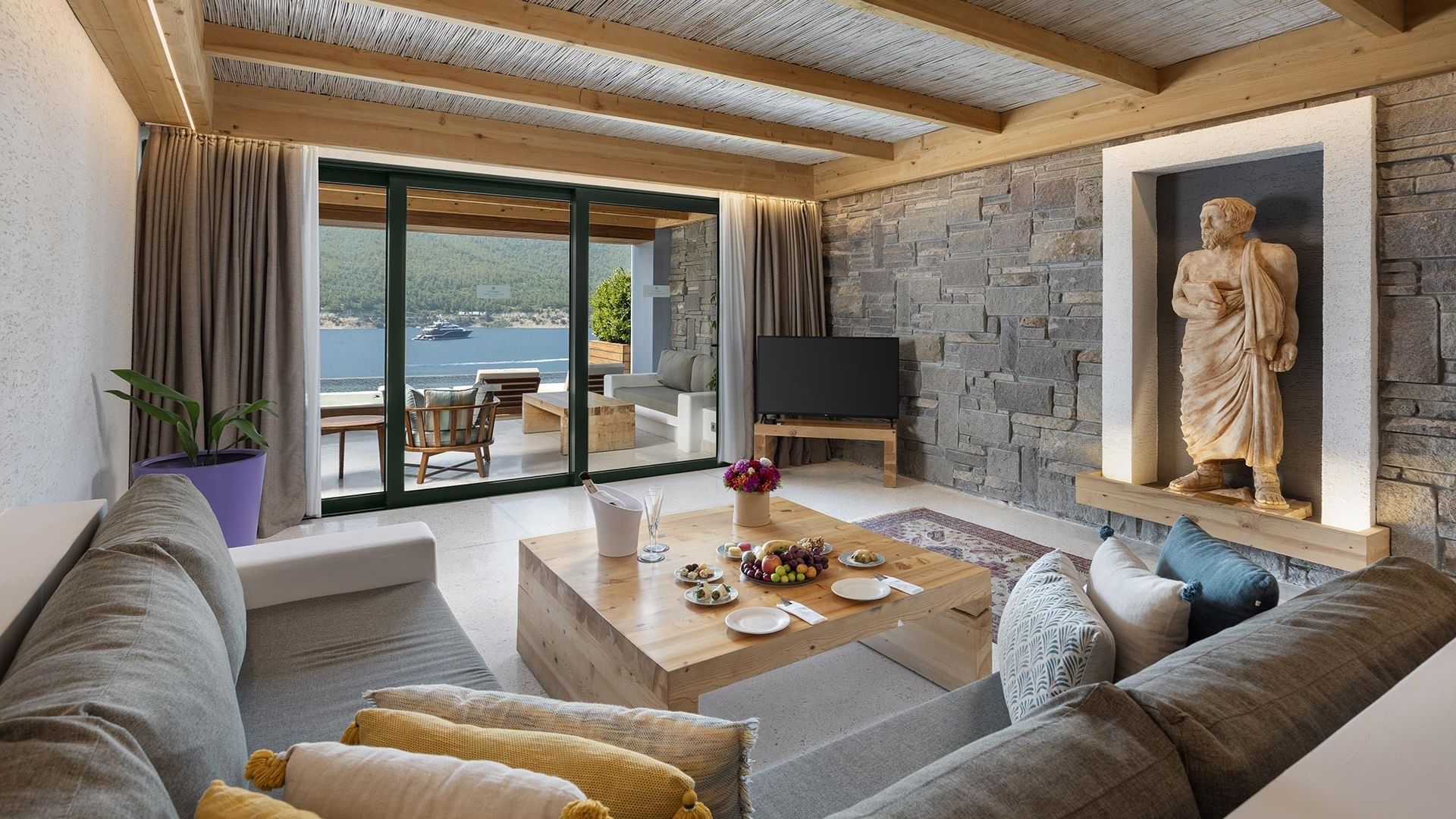 Exclusive Swim-up Room's seating area with couch, coffee table, TV, stone wall, and a statue, overlooking sea and swim-up pool.