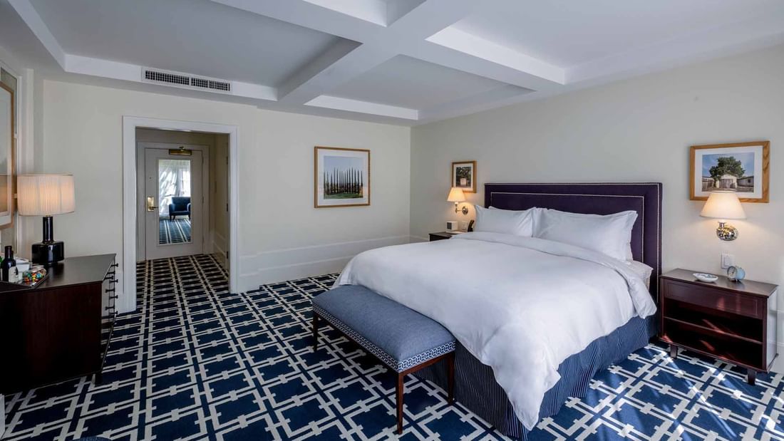 Dresser and ottoman by the cozy bed in Deluxe King Room with carpeted flooring at El Prado Hotel