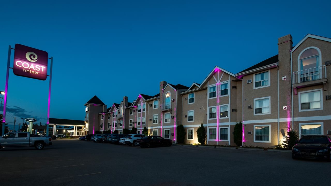 Night view of Coast Hotels with purple and white lighting, several parked cars, and blue sky.